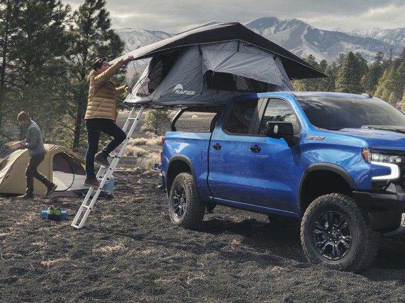 Blue 2026 Chevrolet Silverado 1500 ZR2 with a rooftop tent at an off-road campsite near snow-capped mountains.
