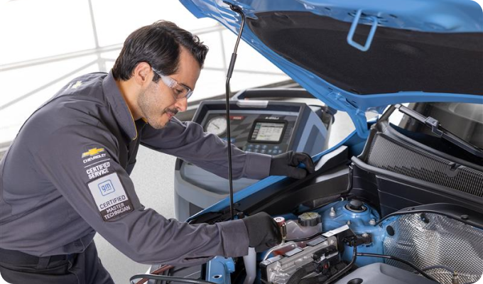 A man in a blue shirt repairs a car, focused on the engine with tools nearby.