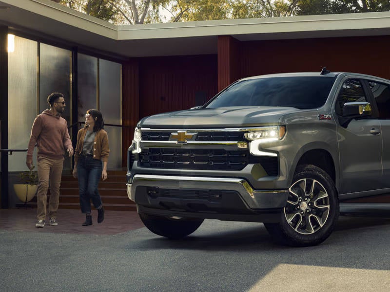 A silver 2026 Chevrolet Silverado 1500 parked in front of a residential house, showcasing its modern design and features.
