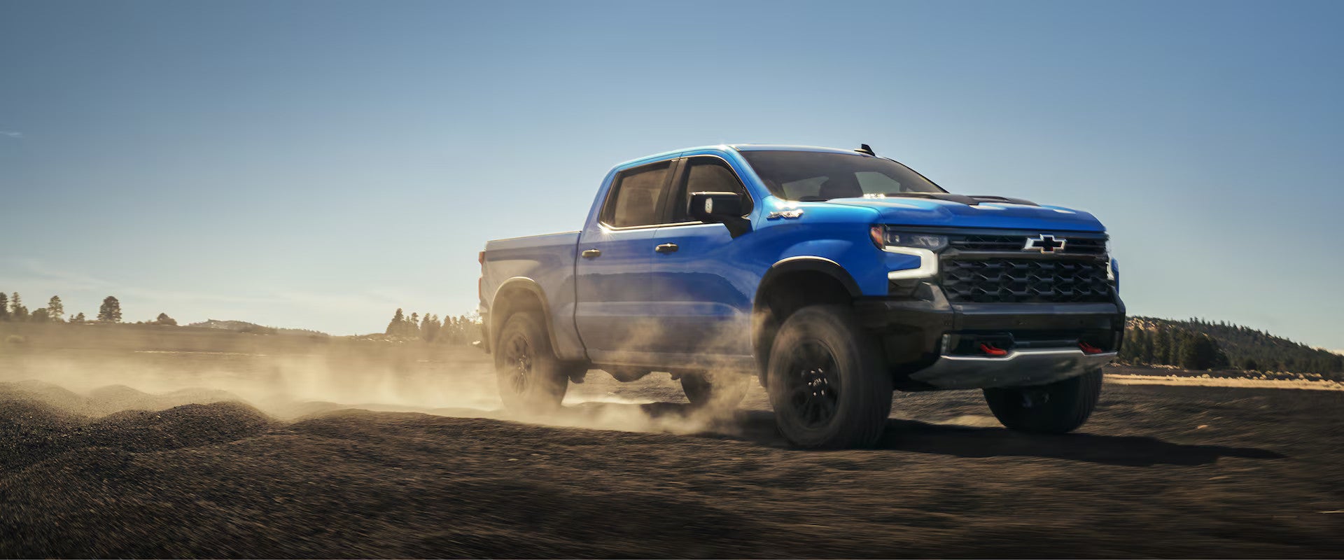 Blue Chevrolet Silverado pickup truck driving off-road on dusty terrain under clear sky.