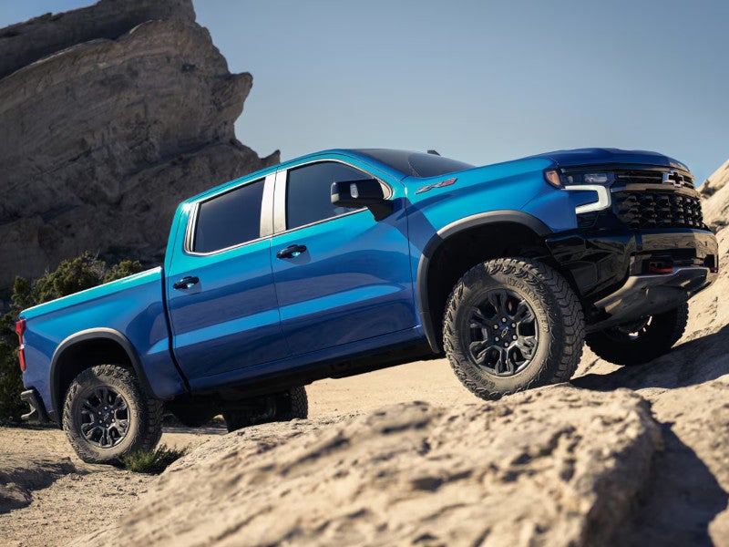 Blue Chevrolet ZR2 pickup truck climbing rocky terrain with large boulders and clear sky background.